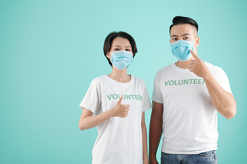Young Asian couple pointing at protective masks and showing thumbs-up when volunteering together