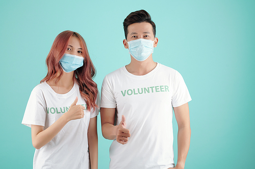 Young Asian students in volunteer t-shirts and medical masks showing thumbs-up and looking at camera