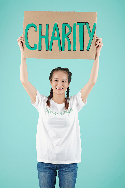 Studio portrait of happy young Asian female volunteer holding charity sign above her head and smiling at camera