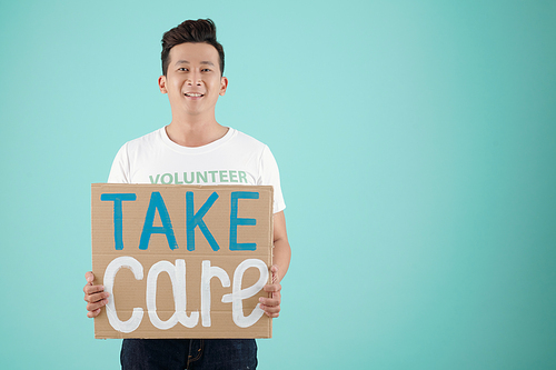 Handosme young Asian volunteer holding cardboard placard with take care inscription and smiling at camera