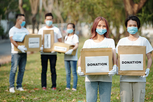 Positive young Asian volunteers in medical masks holding cardboard boxes of food and clothes for people who lost their job due to coronavirus pandemic