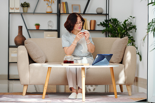 Happy elegant senior Vietnamese woman enjoying black te and watching movie or show episoda on tablet computer