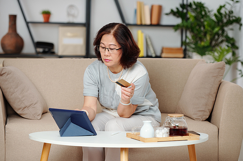 Serious Asian elderly woman in glasses making purchases in online store and paying with credit card