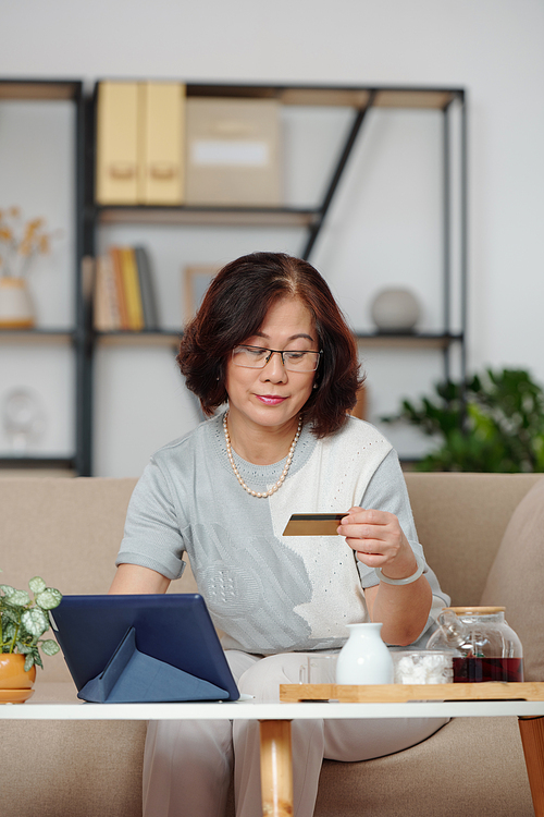 Asian senior woman entering information from credit card when drinking tea and shopping online via tablet computer