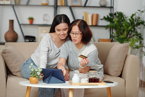 Pretty smiling young Asian woman helping mother to shop online and pay for purchases when they are sitting on sofa in living room and drinking tea