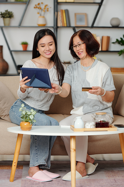 Happy senior Asian woman and her adult daughter enjoying spending time together, they are drinking tea and shopping online