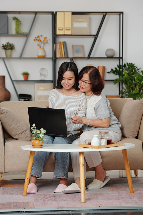 Hugging happy Asian mother and daughter sitting on sofa with laptop and making purchases in online shop