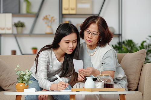 Pretty young Vietnamese woman checking bills and helping mother with home bookkeeping