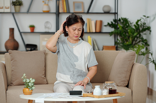 Confused pensive senior Asian woman looking at many bills on coffee table in front of her