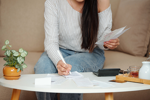 Cropped image of young woman doing home bookkeeping, checking bills and writing in notebook