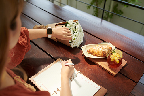 Hands of woman checking notification on her smartwatch and taking notes in planner when eating breakfast in cafe