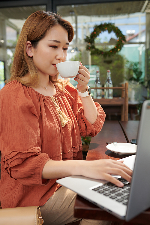 Young woman drinking cup of morning coffee and working on laptop on coffeeshop