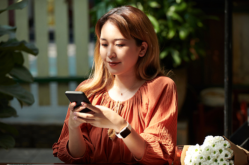 Portrait of positive young woman sitting at table in outdoors cafe and checking news on social media