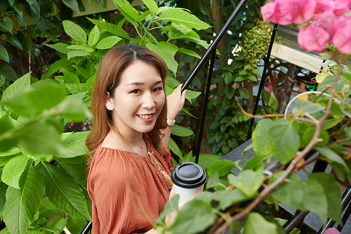Positive young woman enjoying walking in greenhouse and drinking fresh coffee