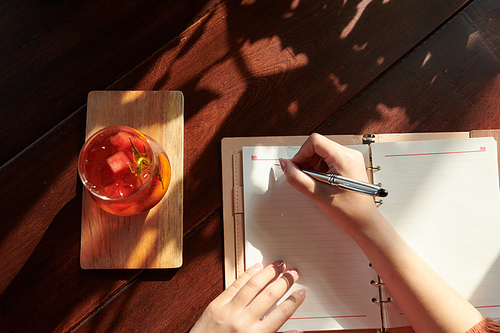 Hands of woman drinking refreshing beverage and writing plans for the day in notepad