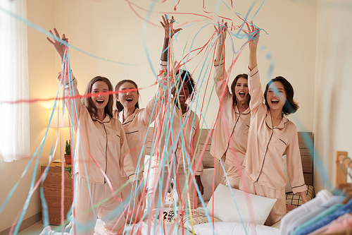 Excited shouting girls in silk pajamas standing on bed and raising arms with plastic ribbons attached to fingers