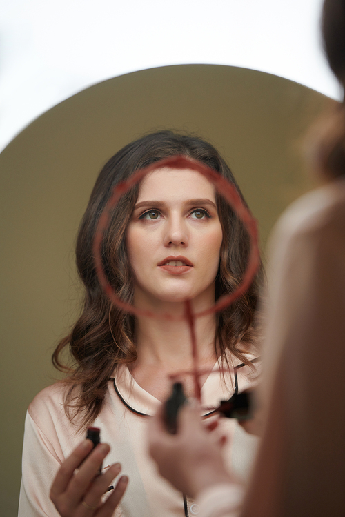 Young woman drawing venus astrology sign with red lipstick on round mirror