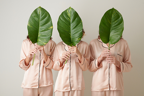 Three young women in light pink silk pajamas holding faces behind big green leaves