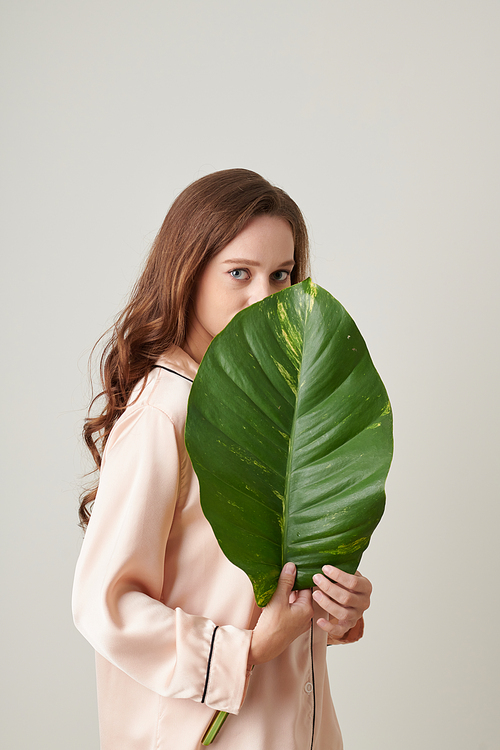 Portrait of serious young woman holding big green leaf and looking at camera