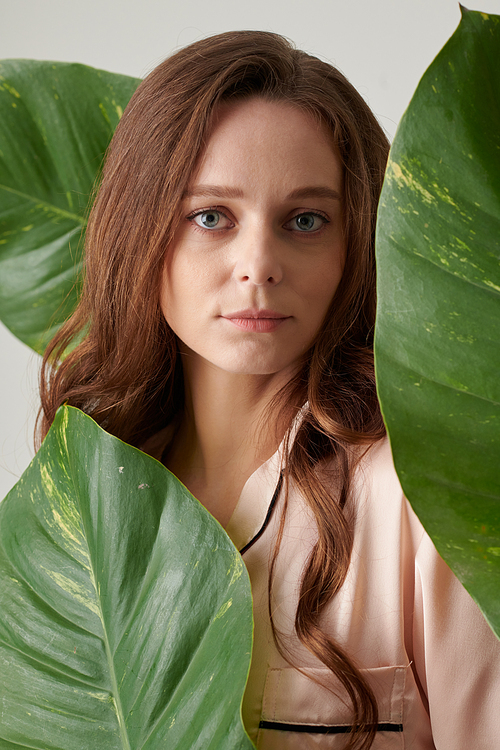 Portrait of unsmiling young brunette with long hair standing among green foliage