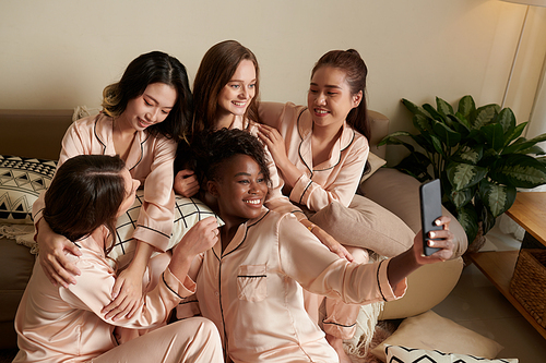 Smiling happy young woman in silk pajamas taking selfie with friends around