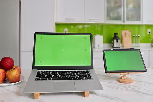 Two wireless mobile gadgets with black crosses on green screens standing on wooden holders on white marble kitchen table