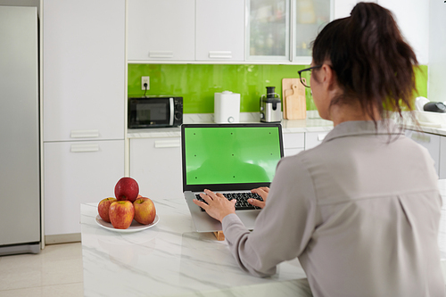 Rear view of young brunette woman typing on keypad of laptop with green screen while working with mobile application at home