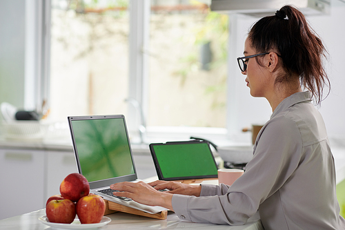 Side view of young modern businesswoman in casualwear typing on laptop keypad with green screen during work in home office