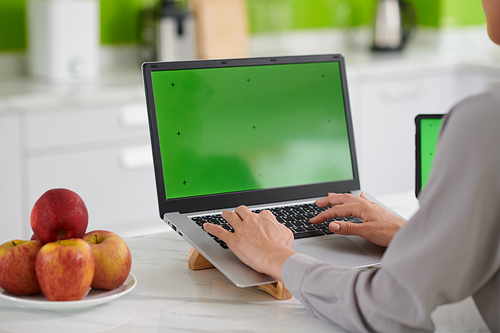 Hands of young businesswoman or freelancer working in front of laptop with green screen while sitting by white marble kitchen table