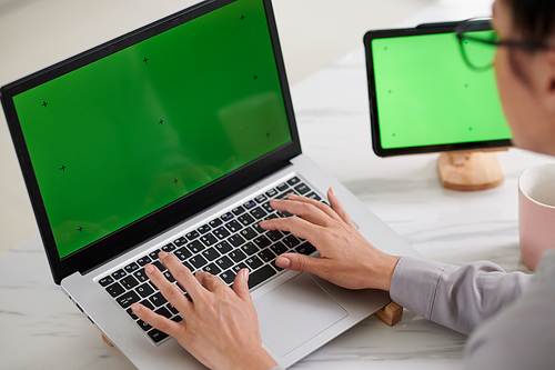 Hands of young woman touching keypad with laptop with green screen while sitting in front of wireless gadget by workplace in home office