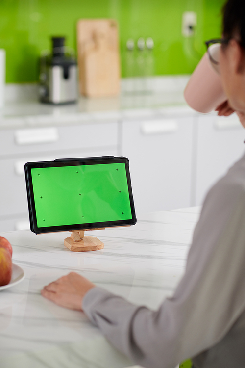 Green screen of tablet on wooden holder and young female home office worker sitting in front of gadget and having tea or coffee