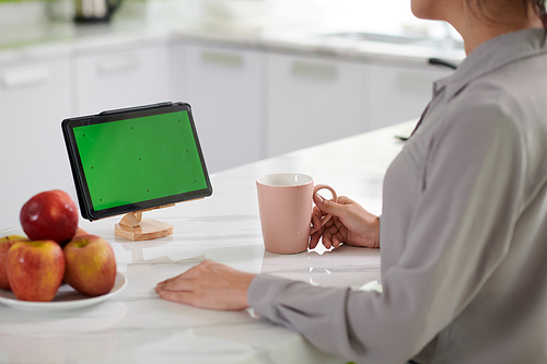 Young female employee with cup of coffee sitting by white marble kitchen table in front of tablet with blank green screen during work