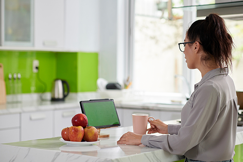 Side view of young woman in casual shirt looking at green screen while sitting in front of tablet by kitchen table, working and having tea