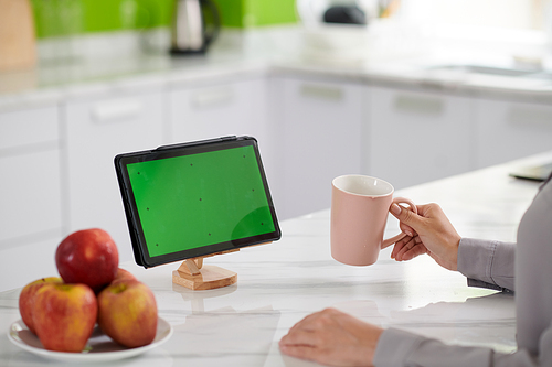 Green screen of tablet standing on wooden holder on white marble kitchen table where young home office worker sitting