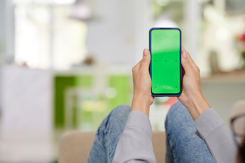 Smartphone with blank green screen held by young woman in casualwear sitting in front of camera and using new mobile application