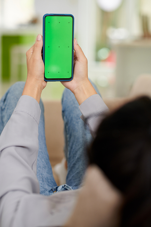 Blank green screen of smartphone in hands of young woman in casualwear sitting in front of camera and using application