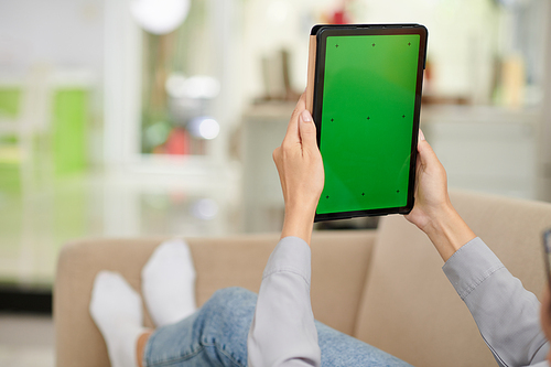 Close-up of young modern woman in casualwear holding tablet with blank green screen while relaxing on couch in front of camera