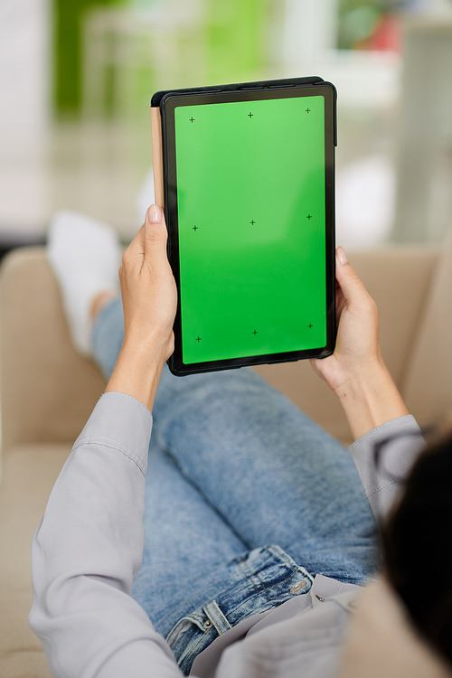 Digital tablet with blank green screen in hands of young restful woman relaxing on couch in front of camera in home environment