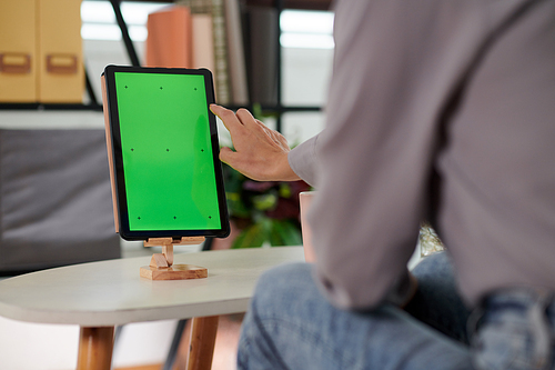 Hand of young woman going to touch blank green screen of tablet on wooden holder while sitting in front of mobile gadget