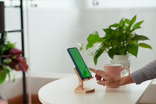 Side view of hand of young businesswoman pointing at green screen of smartphone on wooden holder while going to start new application