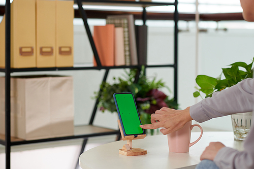 Hand of girl sitting by workplace in home office and pointing at smartphone blank green screen while starting new mobile application