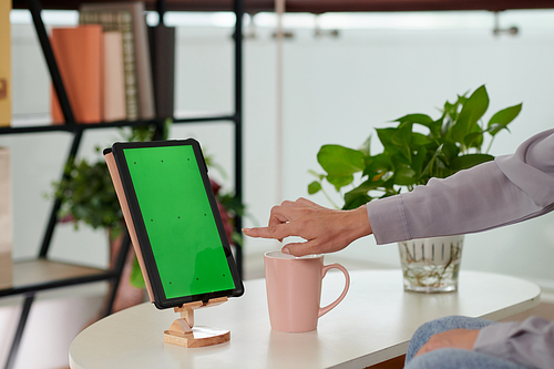 Hand of young female home office worker pointing at green screen of tablet on holder while making presentation of mobile application