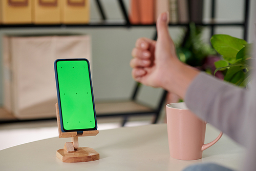 Focus on smartphone with blank green screen on wooden holder standing on workplace of home office worker keeping thumb up