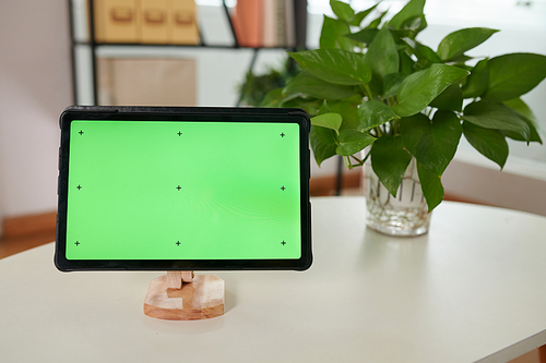 Digital tablet with blank green screen on wooden holder standing on white table which is workplace of modern home office worker