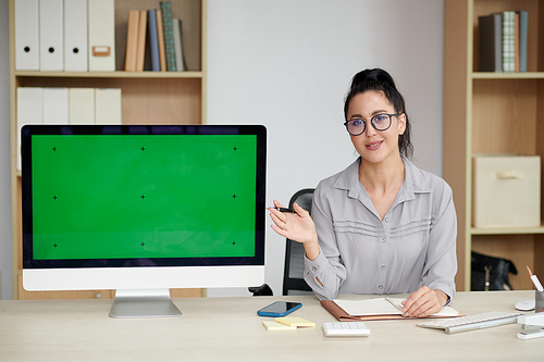 Young brunette businesswoman in eyeglasses and casualwear making presentation of new application while pointing at blank green screen