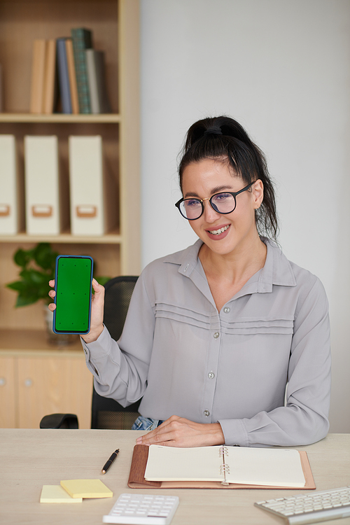 Happy young elegant businesswoman holding smartphone with blank green screen while making presentation of new mobile application
