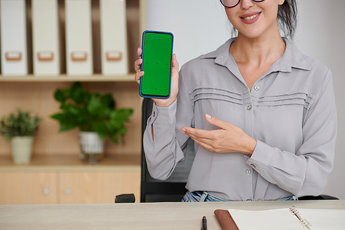 Close-up of young smiling woman presenting new mobile application in smartphone with blank green screen while sitting by workplace