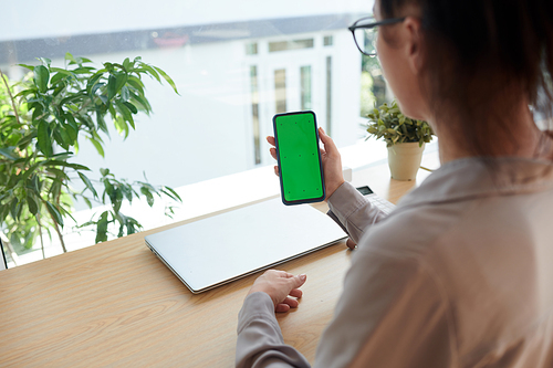 Hand of young businesswoman holding smartphone with blank green screen over workplace while using new mobile application