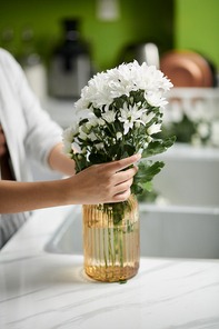 Closeup image of woman putting blooming flowers in vase on kitchen counter