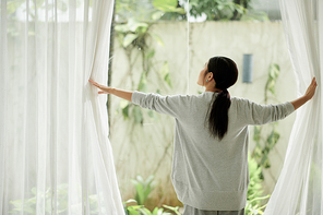 Young woman opening curtains of big window, view from back